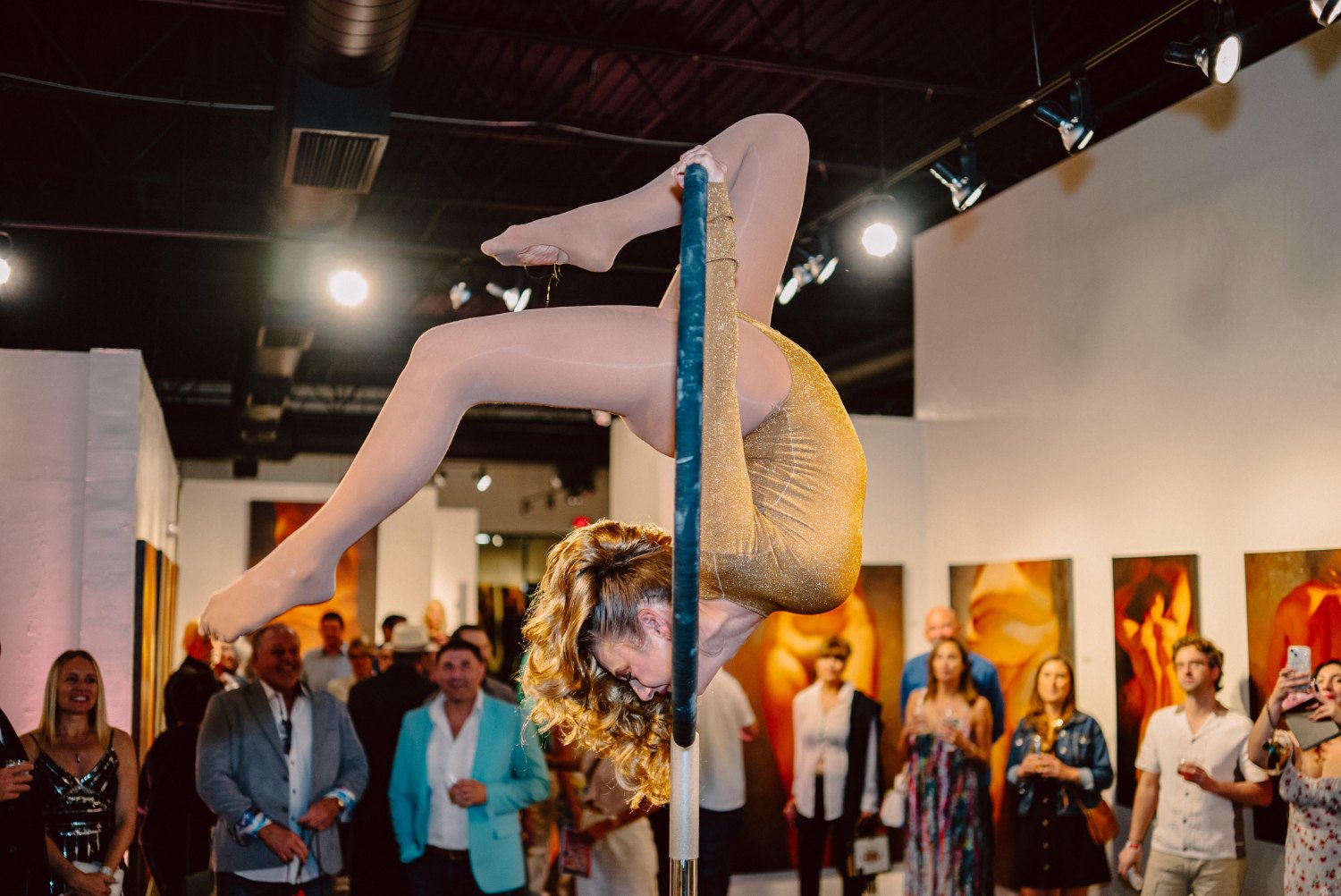 Aerialist in a gold costume performing on a pole as guests watch in the gallery